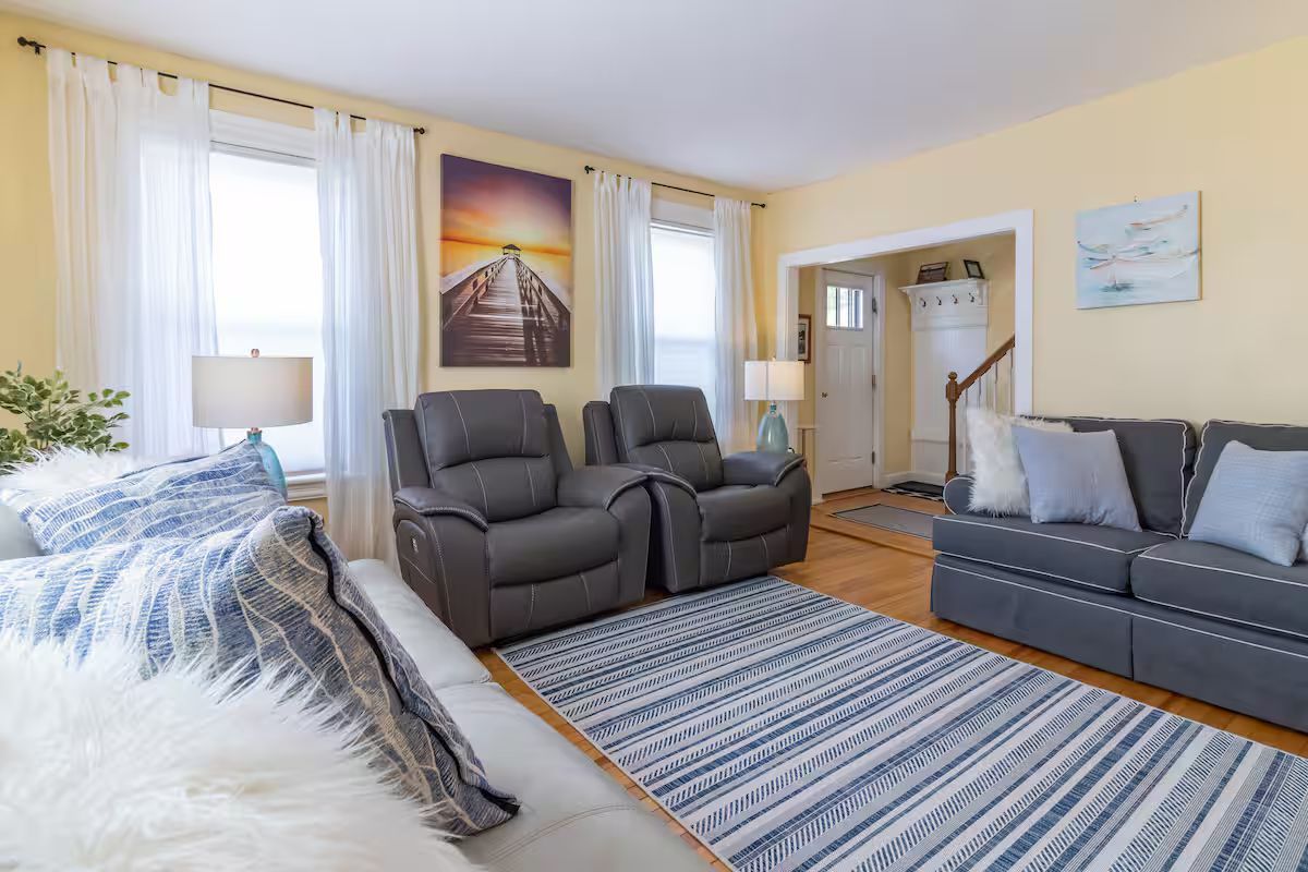 Bright living room with blue-gray sofas, recliners, striped rug, and wall art by a staircase