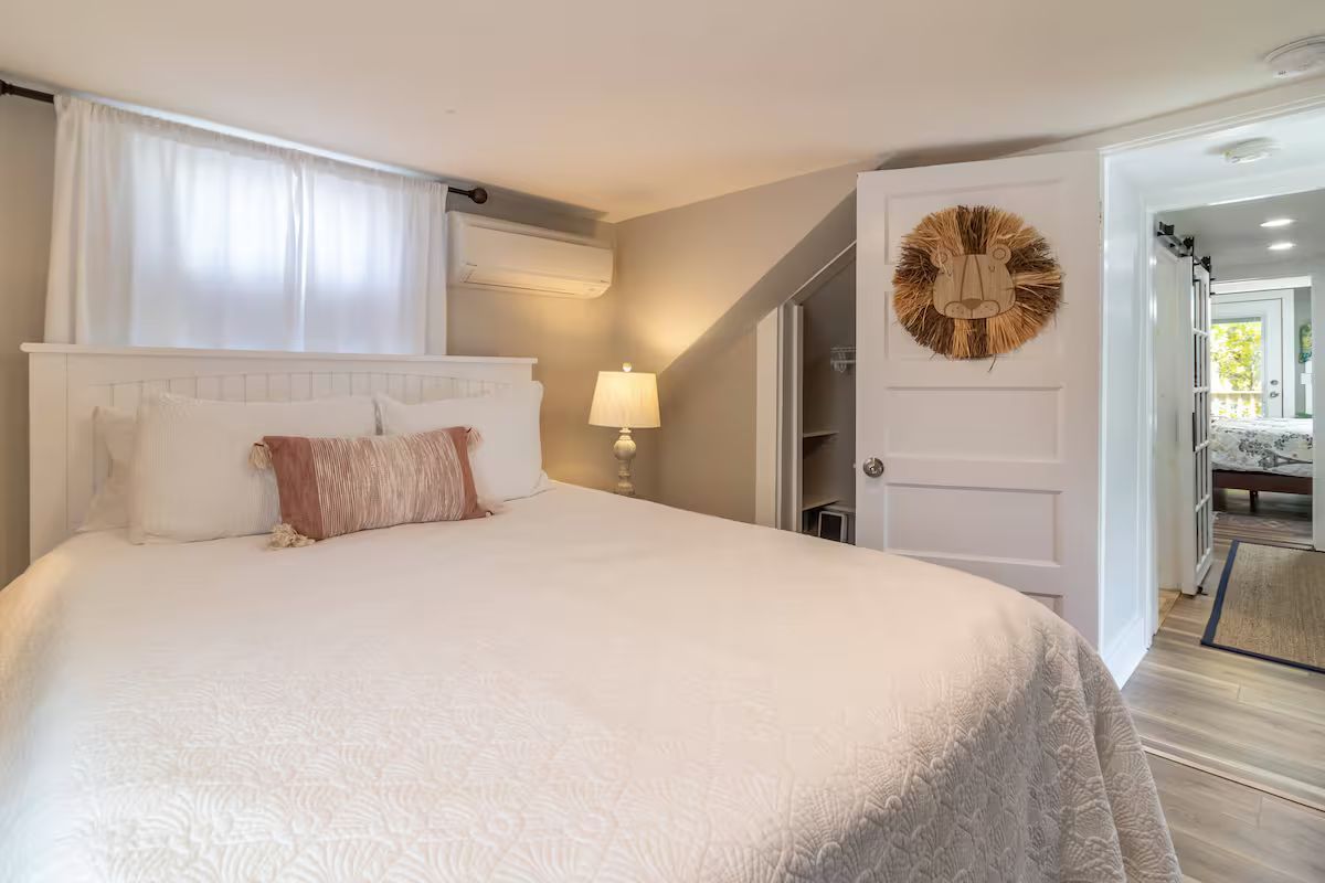 Bright bedroom with white bed, soft pink pillows, lamp, and open doorway to a hallway