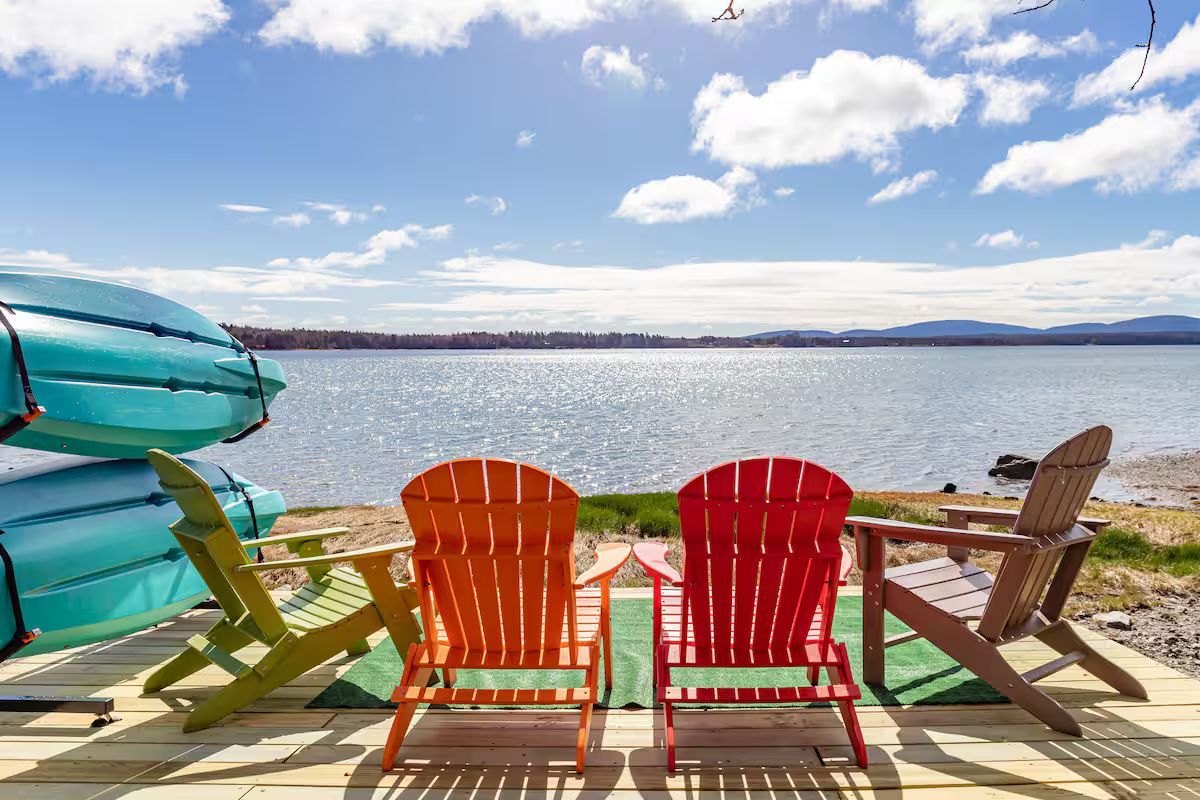 Colorful Adirondack chairs on a sunny lakeside deck with kayaks and a calm water view