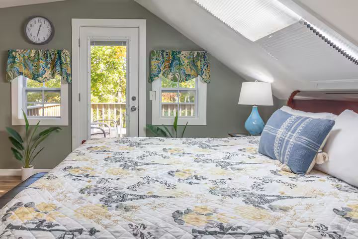 Bright attic bedroom with a floral bedspread, skylights, a door to a balcony, and a blue lamp.