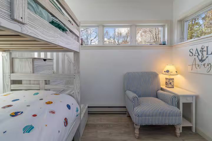 Bright child’s bedroom with white bunk bed, striped armchair, small table, and high window.