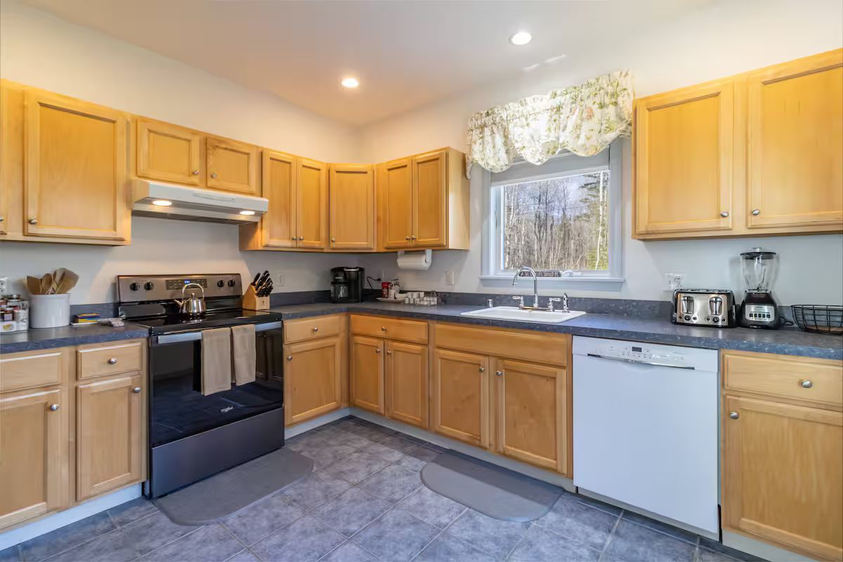Bright kitchen with light wood cabinets, black stove, white dishwasher, and a window above the sink
