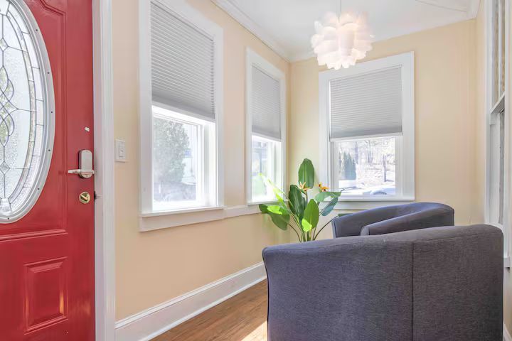 Bright entryway with red door, beige walls, windows, and a gray chair near a potted plant.