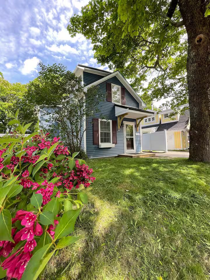 Blue house with white trim, pink flowers in front yard, and a sunny green lawn under a cloudy sky