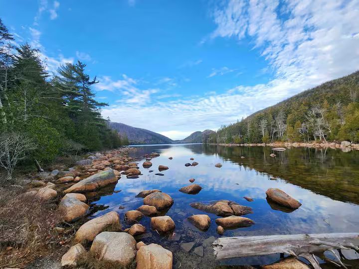 Rocky river shoreline reflecting evergreen hills and blue sky on a calm day