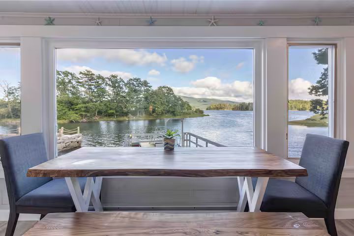 Dining table and chairs overlooking a calm lake and trees through large windows