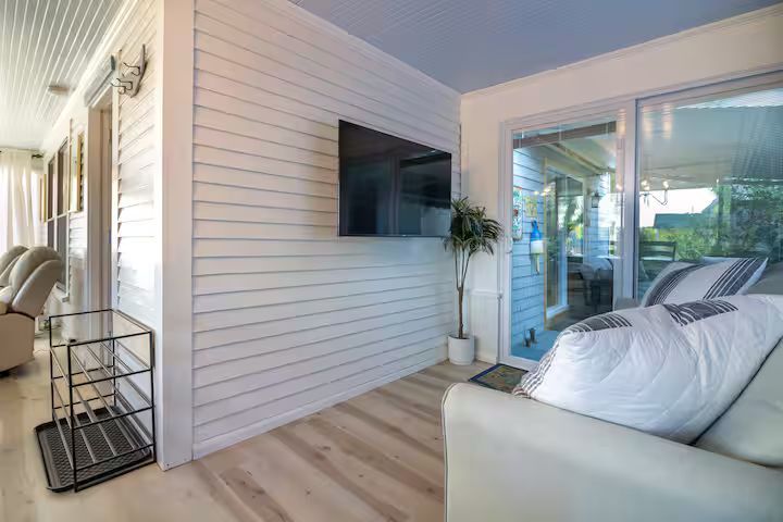 Modern living room with wall-mounted TV, light wood floors, white sofa, and large windows.