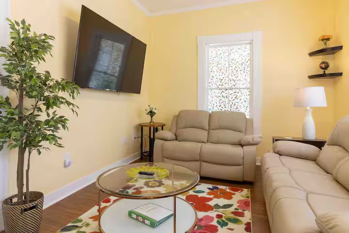 Cozy living room with beige sofas, wall-mounted TV, glass coffee table, colorful rug, and tall plant