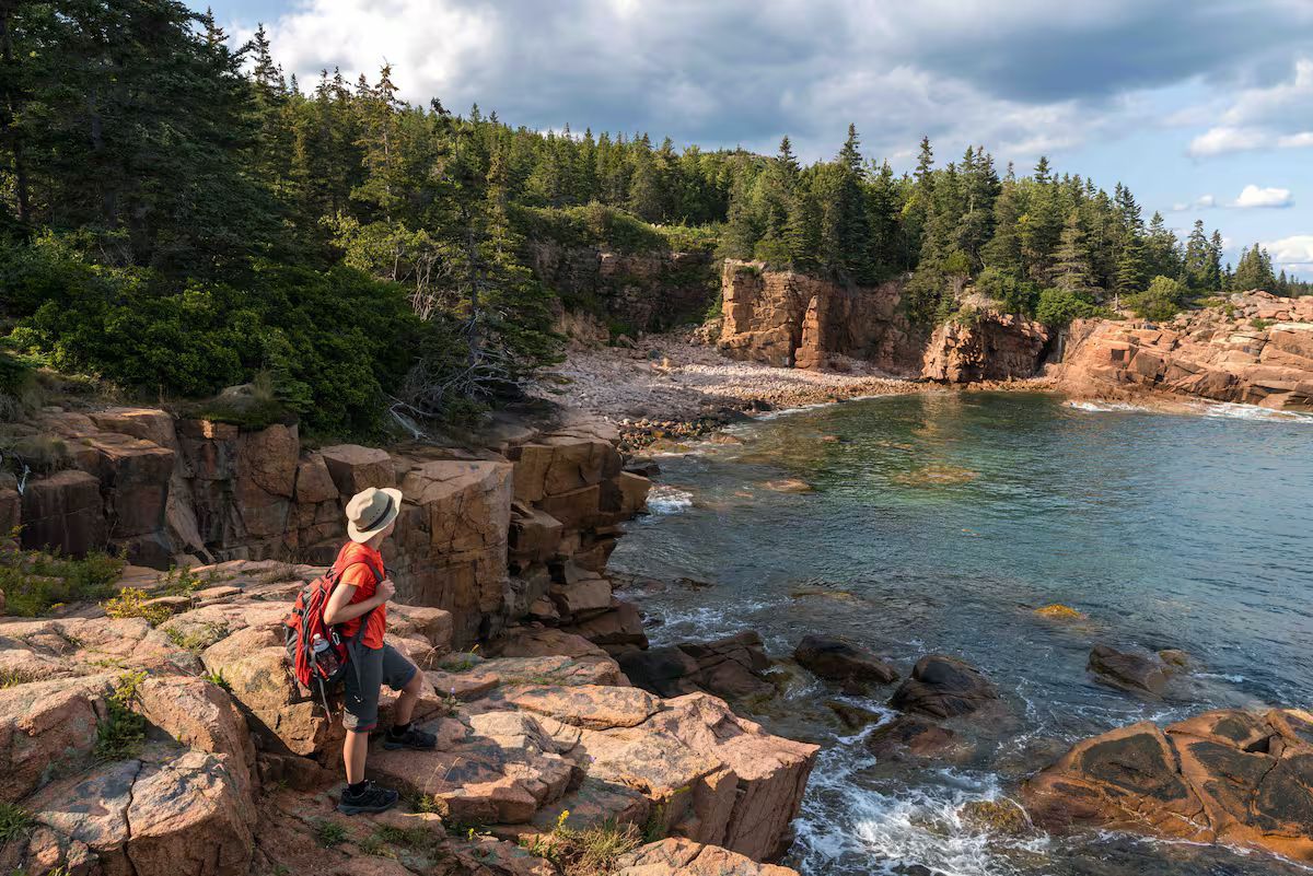 Hiker with red backpack on rocky shoreline overlooking blue water and forested cliffs