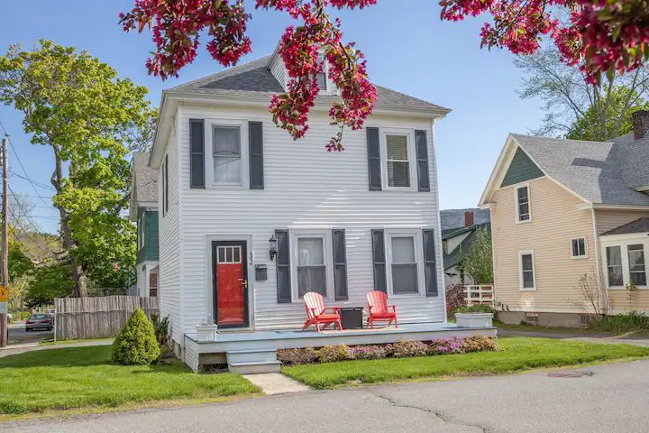 White two-story house with red door and chairs on a small front porch, framed by flowering trees