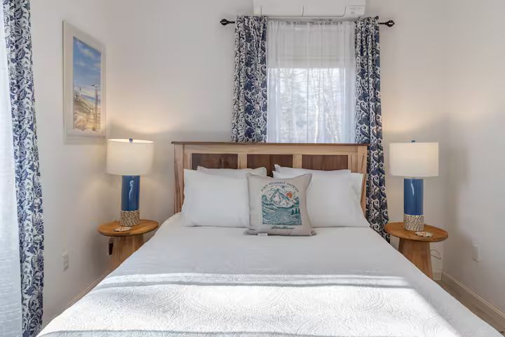 Bright bedroom with a wooden bed, white bedding, blue lamps, and floral curtains at the window