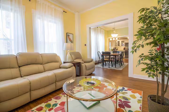 Bright living room with beige sofa, glass coffee table, floral rug, and view into a dining room