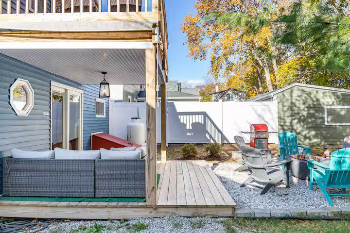 Backyard patio with gray sectional, wood deck, red chairs, and autumn trees beside a house.