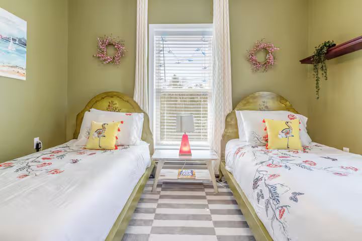 Twin beds in a pastel green bedroom with a window, floral pillows, and a striped rug between them