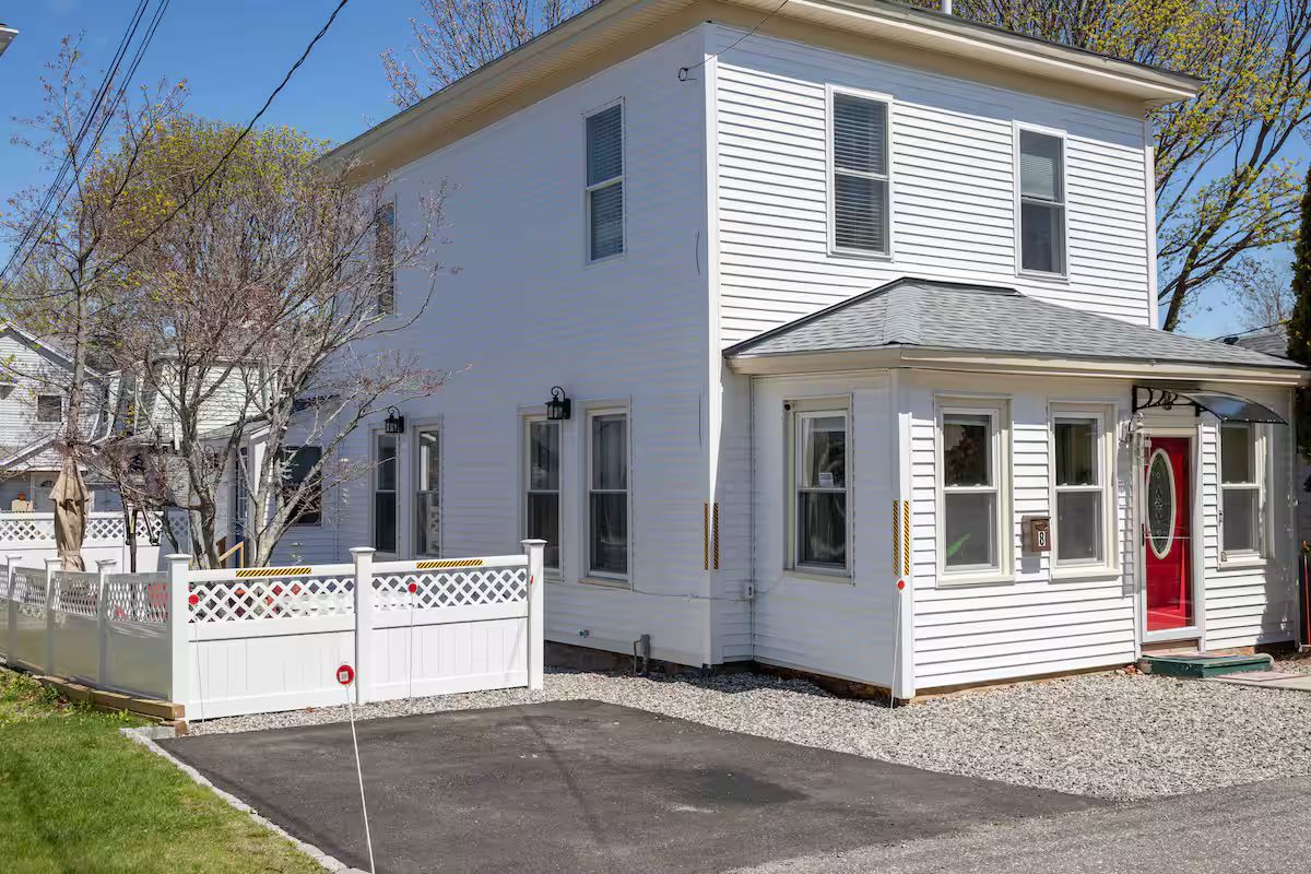 White two-story house with a red front door and fenced yard on a sunny street