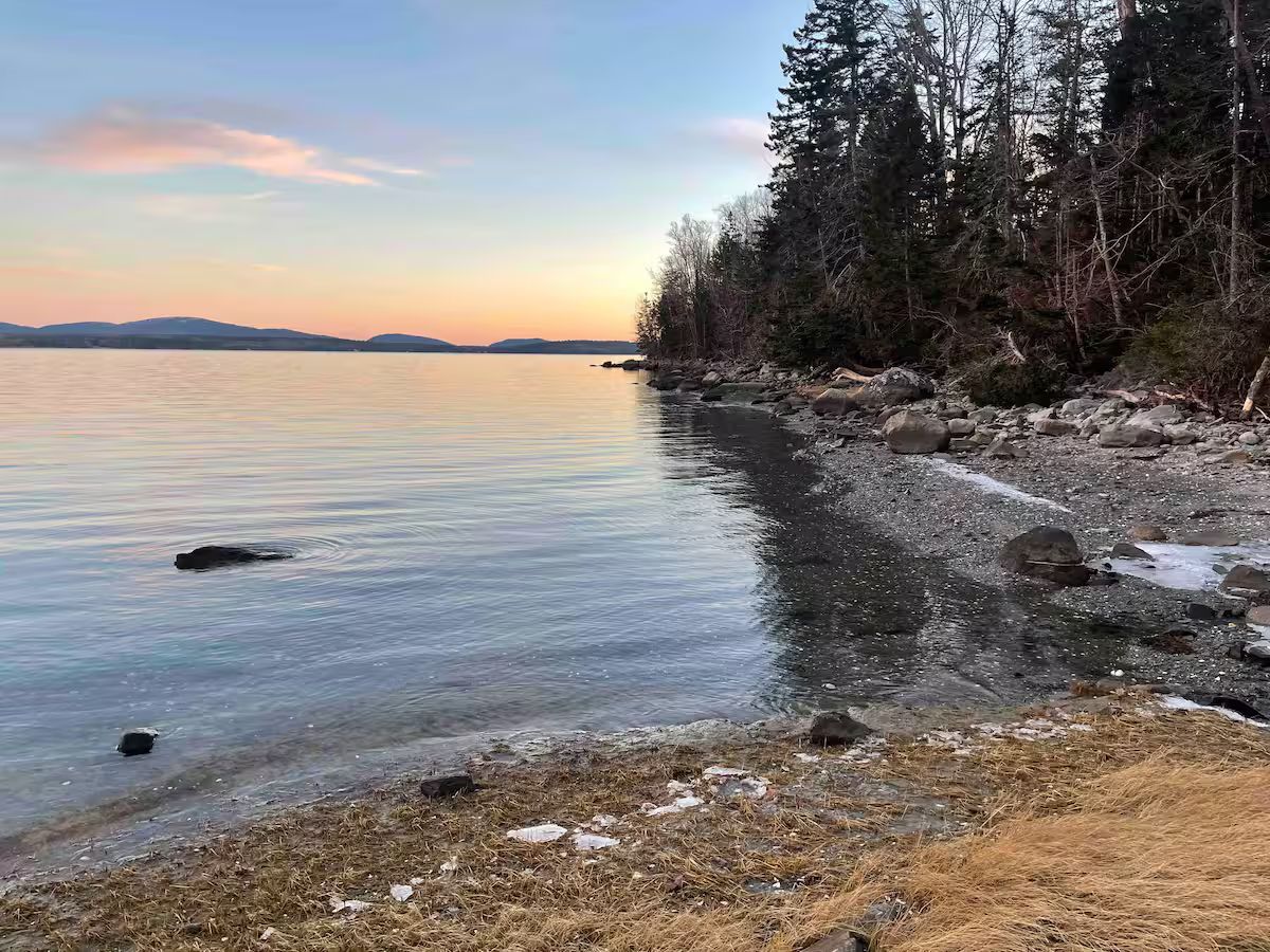 Rocky lakeshore at sunset with calm water, distant islands, and evergreen trees along the shore