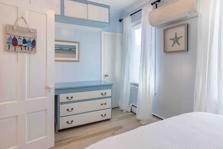 Bright blue-and-white bedroom with dresser, wall art, and sheer curtains beside a bed