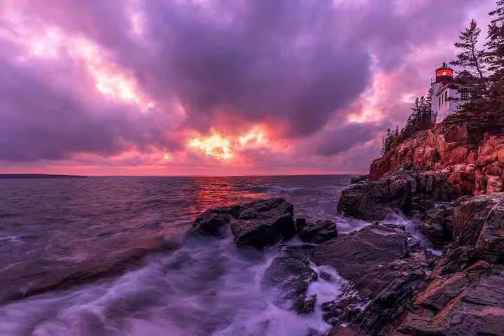 Purple sunset over a rocky shoreline with waves crashing below a cliffside lighthouse