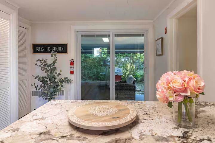 Bright kitchen with granite island, tray, pink flowers, and sliding glass door to a leafy patio.