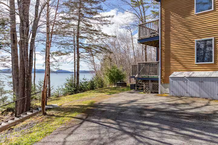 Lakefront home with trees, driveway, and a view of water under a bright sky