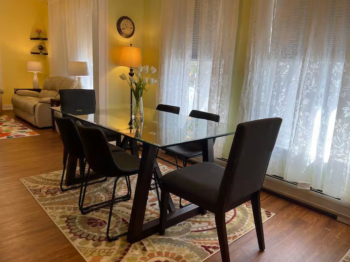 Dining area with black glass table, four chairs, rug, and sheer curtains beside a cozy living room