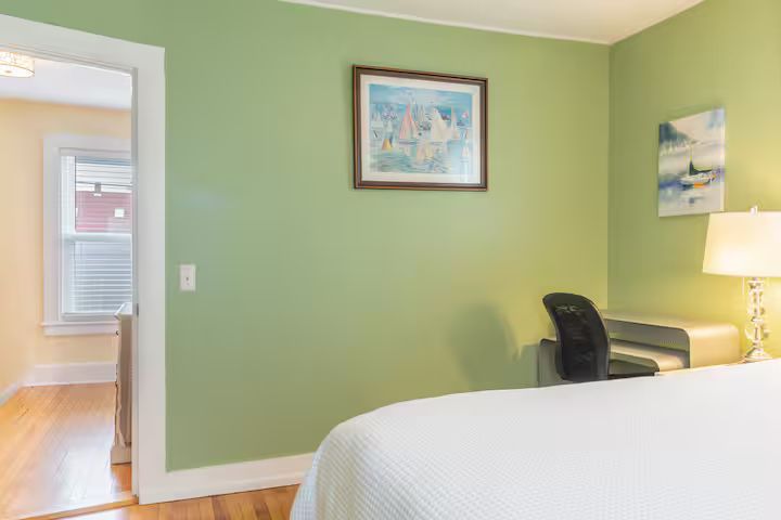 Green bedroom with white bed, black chair, framed art, and doorway to a hall with hardwood floors