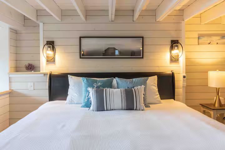 Cozy bedroom with a king bed, white shiplap walls, blue pillows, wall sconces, and framed art above the headboard