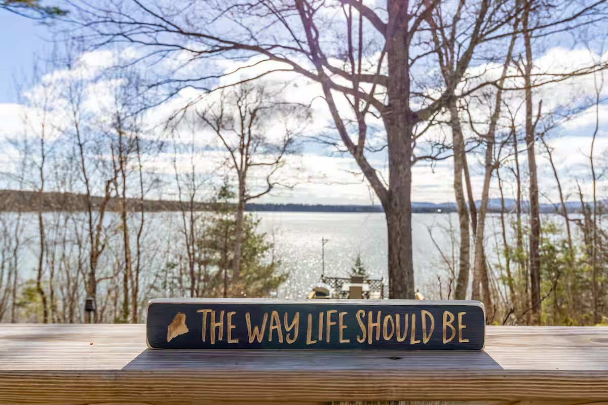 Sign on wooden deck reading “THE WAY LIFE SHOULD BE” overlooking a lake and bare trees