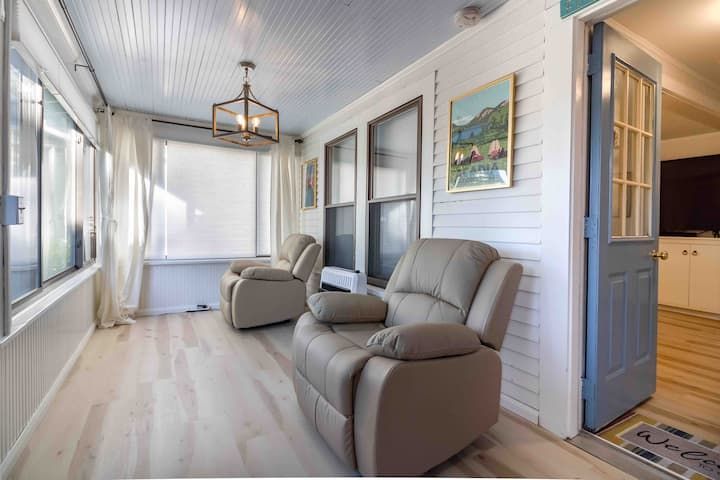 Bright enclosed porch with two gray recliners, white shiplap walls, and blue-framed door leading to a kitchen