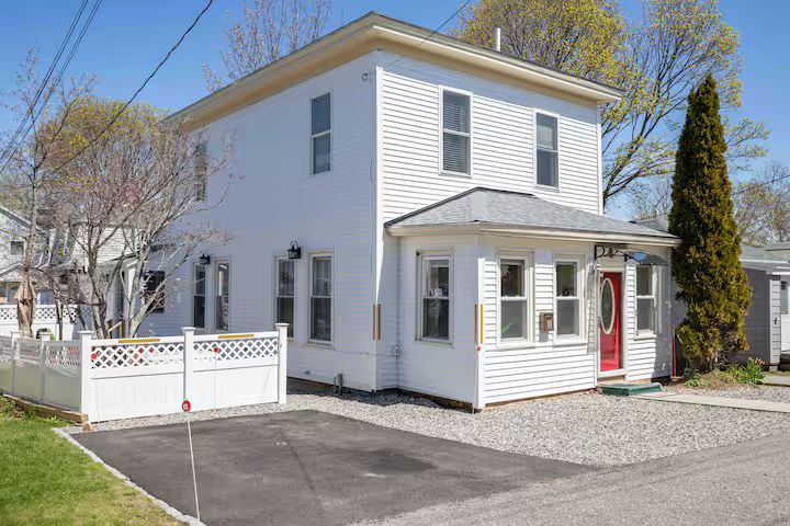 White two-story house with red front door, white fence, and gravel driveway on a sunny day