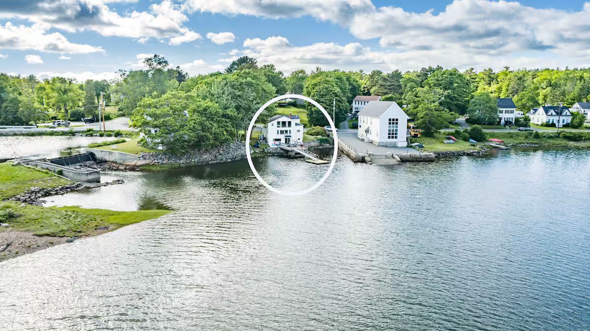Calm lakeside view with a small white house, dock, and trees under a partly cloudy sky