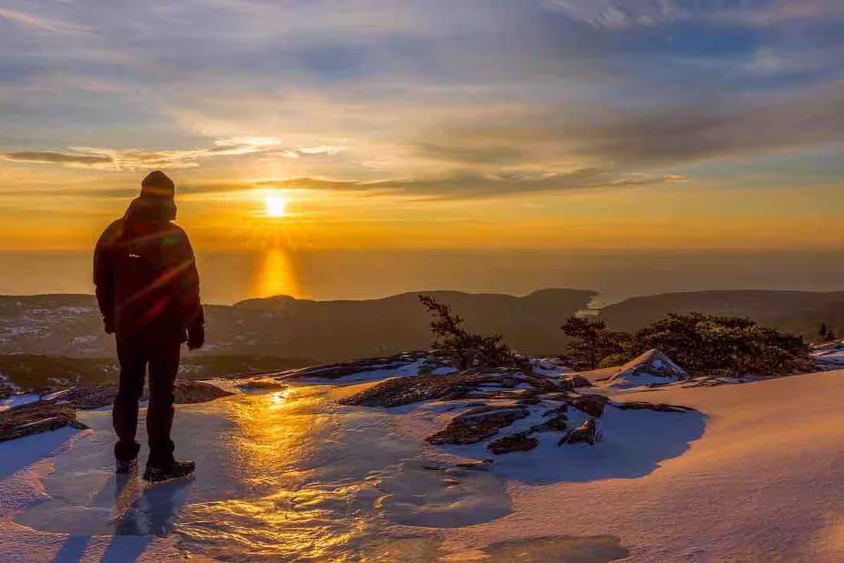 Person standing on a snowy ridge at sunset, overlooking a glowing golden horizon
