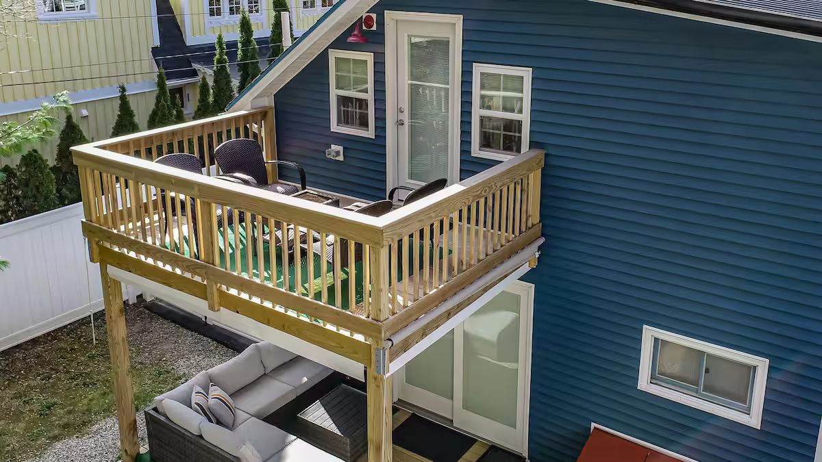 Wooden balcony deck attached to a blue house, with patio furniture and a white fence below.