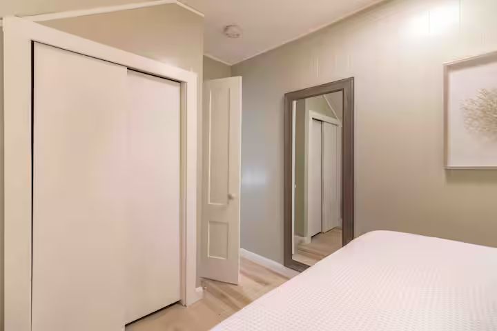 Bright bedroom with beige walls, white closet doors, a tall mirror, and an open doorway to a hallway