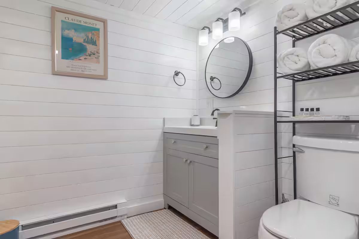 Bright white bathroom with vanity, round mirror, shelving, and toilet.