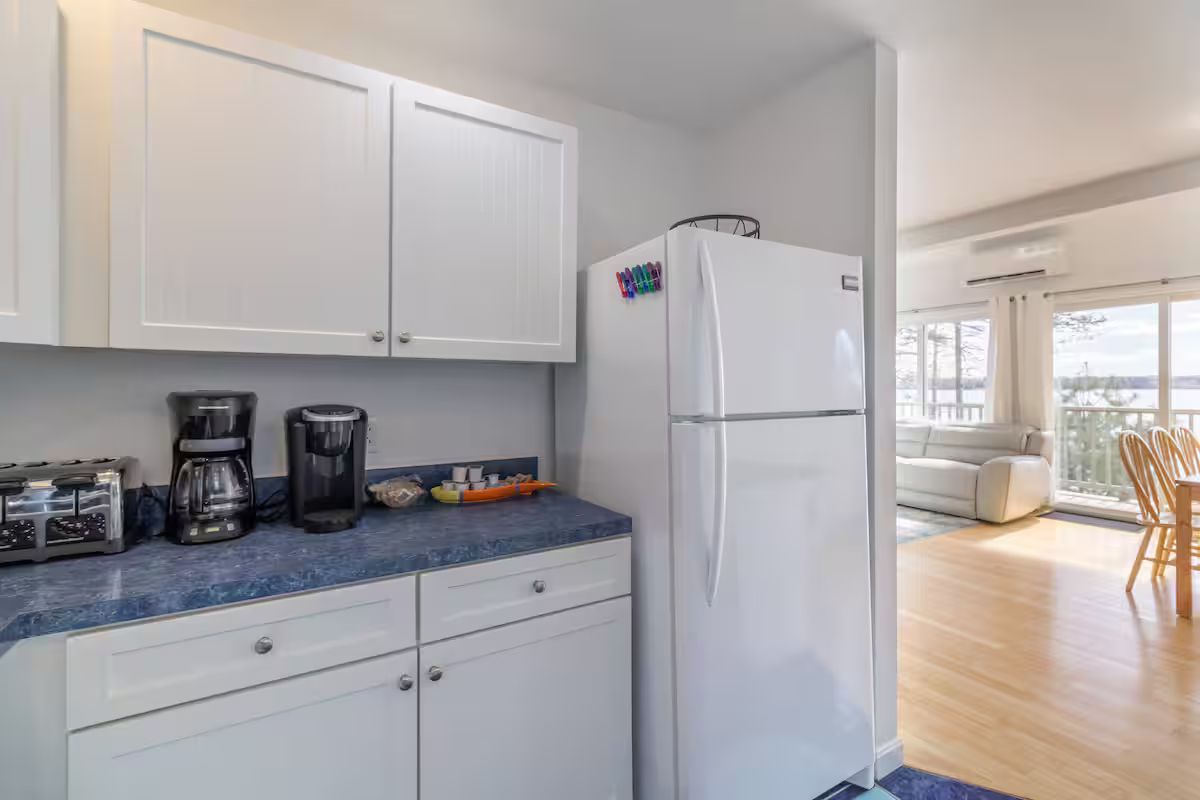 Compact white kitchen with blue countertop, coffee makers, and a white refrigerator opening to a bright dining area