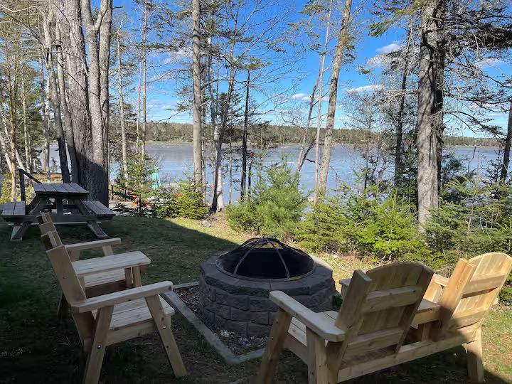 Wooden chairs around a fire pit overlooking a lake through trees on a sunny day