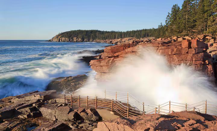Waves crash against red rocky cliffs at a coastal lookout, sending white spray over the shore.