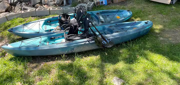 Two blue kayaks on grass, one stacked on top of the other with fishing gear attached.