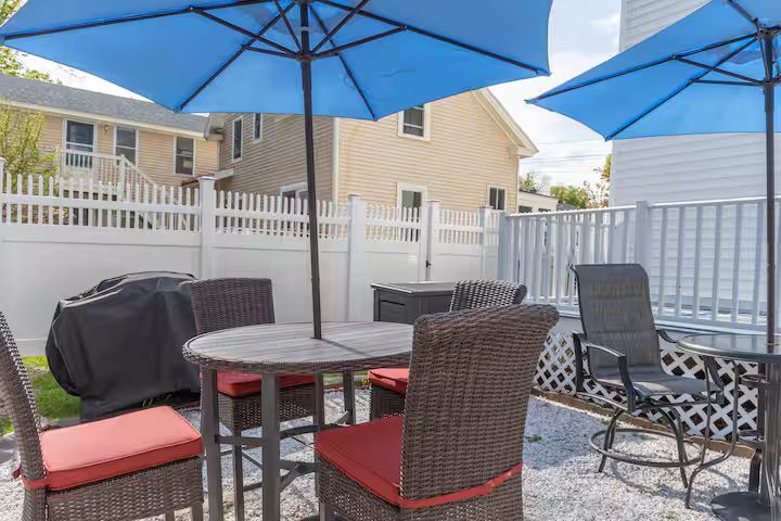 Back patio with blue umbrellas, wicker chairs, table, and grill on a gravel surface