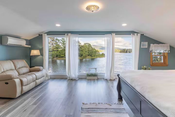 Bright bedroom with a lake view through large windows, gray flooring, recliner, and a bed in the foreground