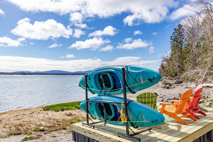 Kayaks stacked by a lakeshore with orange chairs and trees under a blue sky