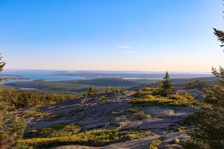 Rocky overlook above a forested valley and distant water under a clear blue sky