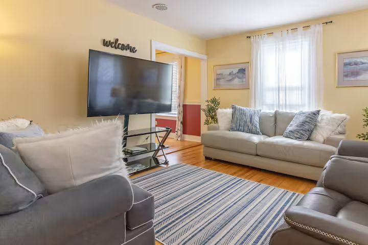 Bright living room with gray sofas, wall-mounted TV, striped rug, and sunlit window.