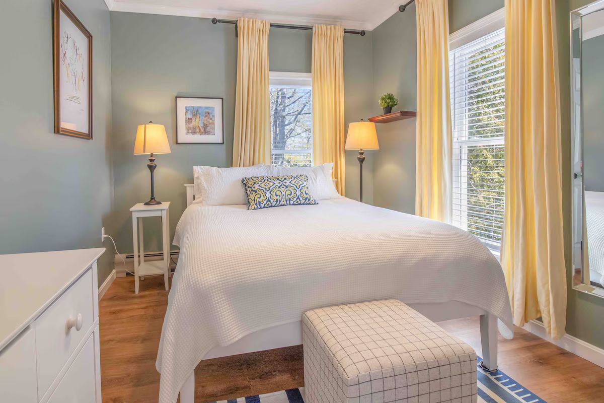 Bright bedroom with a white bed, yellow curtains, bedside lamps, and a window seat by the window