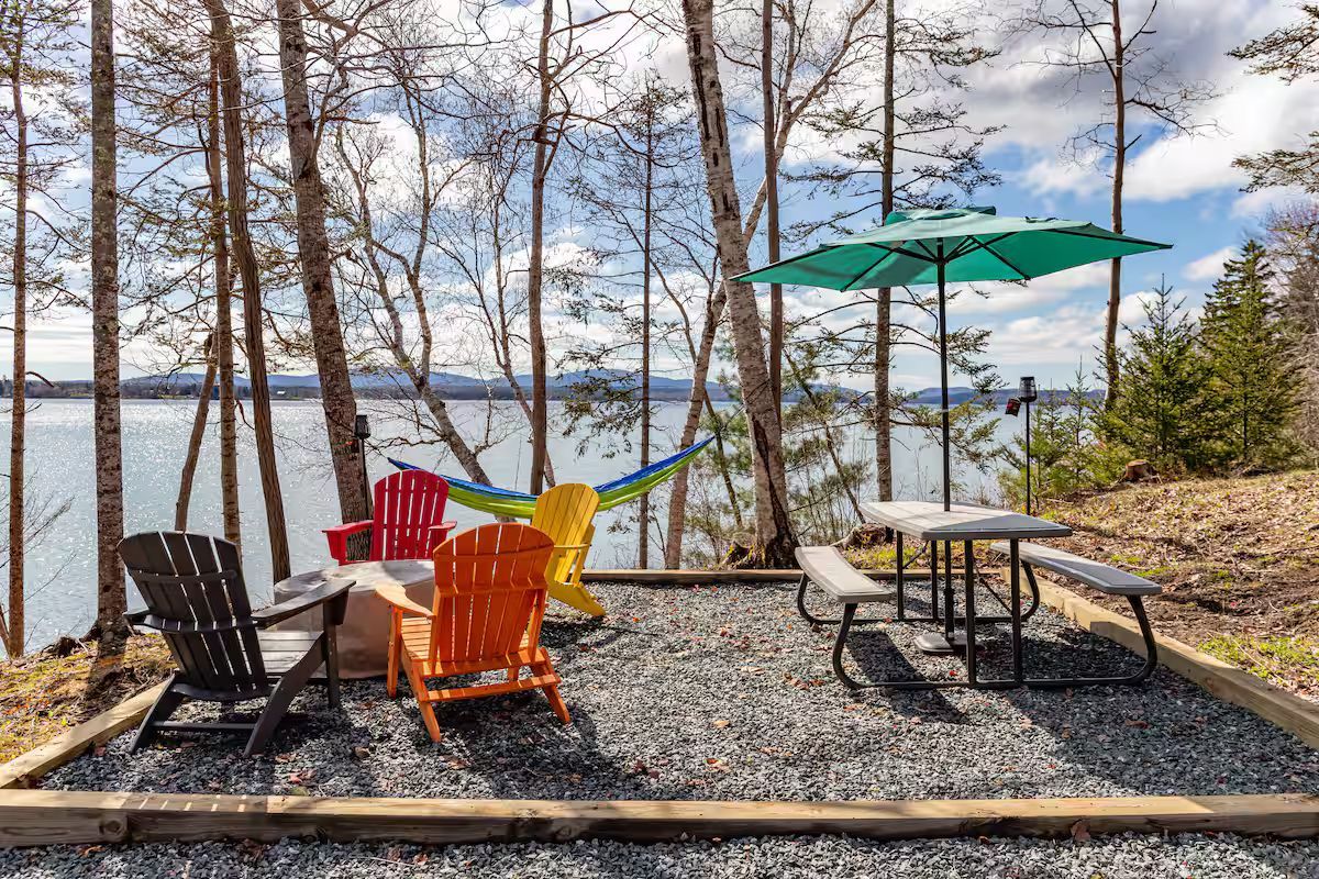 Lakeside patio with colorful chairs, table, and green umbrella among birch trees and gravel seating area