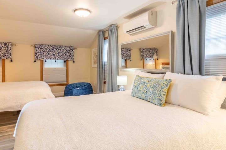 Bright bedroom with two white beds, blue accents, windows with curtains, and a wall-mounted air conditioner
