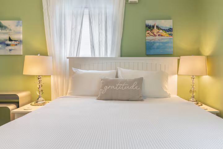 Bright white bed in a pastel green bedroom with bedside lamps, sheer curtains, and coastal wall art