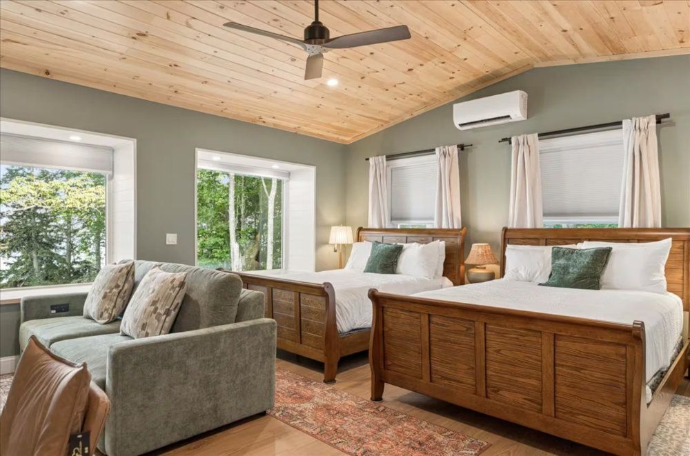 Bright bedroom with two wooden beds, gray chair, wood ceiling, and large windows with curtains