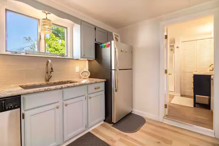 Bright kitchen with stainless-steel fridge, white cabinets, sink by window, and light wood floor leading to hallway